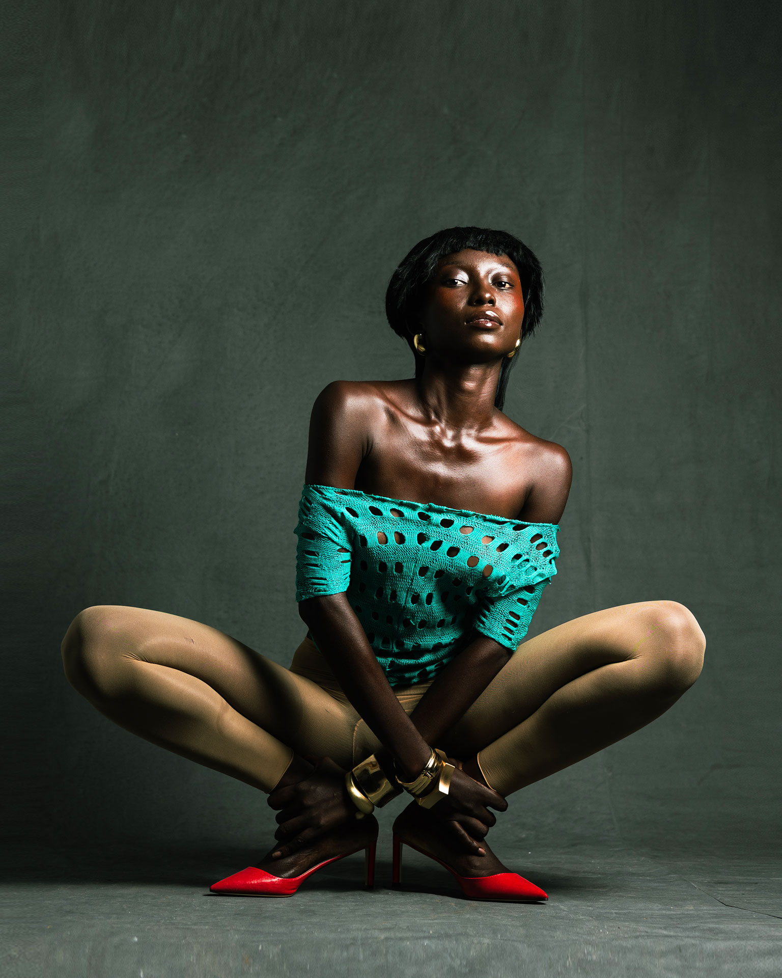 Seated studio pose in turquoise styling and tan tights, with red heels