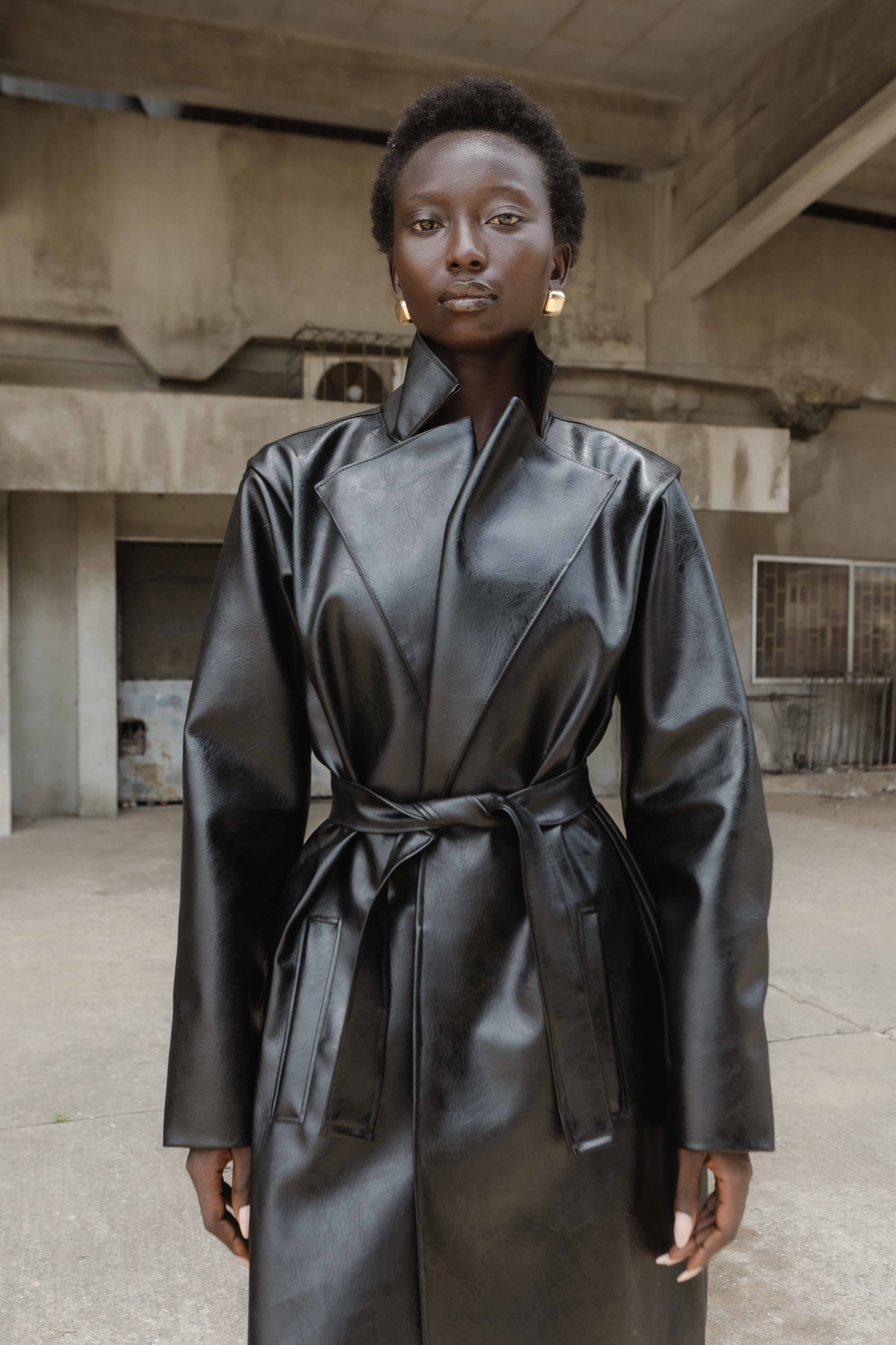 Full-length colour portrait in a belted black coat and grey skirt against bare concrete